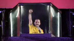 紫と赤のステージにコの字型にガラスが立てられ、その中で、黄色いサッカーユニフォームを着た男性が右手のこぶしを上げて聴衆にアピールしている。左手にはマイクを持っている