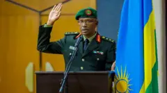 Innocent Kabandana yambaye ikositimu ya gisirikare y'ibara ry'icyatsi kibisi, ishati, karuvati n'ingofero y'icyatsi kibisi ihengetse. Ukuboko kumwe gufashe ku ibendera ry'u Rwanda, mu gihe ikiganza cy'ukundi kuboko kizamuye. Ahagaze imbere y'indangururamajwi n'ameza maremare yo kuvugiraho ijambo. Inyuma ye hari urukuta rw'inzu. Ifoto yo mu mwaka wa 2019. 