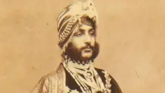 A portrait of Duleep Singh on paper that has aged over time and gone brown. He looks away from the camera and is expressionless. He has a 19th Century traditional Indian turban on as well as clothing. He also has a dark and thick beard.