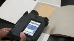 Aparelho preto sendo segurado em cima de um papel com anotações. No visor aparecem alguns números