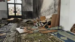 نمای داخل مسجد امام علی پس از انفجار. دیوارهای سیاه و سوخته، پنجره‌های شکسته و لکه‌های خون روی فرش دیده می‌شود. قفسه کتاب یا قرآنی دیده می‌شود که از هم پاشیده و کتاب‌ها به زمین افتاده اند. پارچه‌های شیشه نیز به هر سو پاشیده شده اند. 