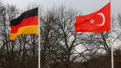 Türkiye ve Almanya bayrakları yanyana gönderde arka planda yapraksız bir ağaç var. 