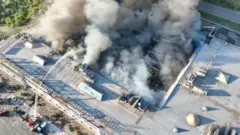 Вид згори на велику промислову або складську будівлю, охоплену сильною пожежею. З даху піднімаються густі клуби чорного та сірого диму. Пожежники з автодрабини спрямовують потужний струмінь води на осередок займання.