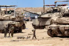 Un soldado israelí camina entre tres tanques de guerra en el desierto, mientras otros tres soldados están parados en un grupo hablando y riendo.