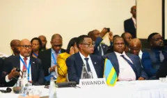 Minisitiri w'u Rwanda James Kabarebe yicaye imbere hagati, yambaye ikositimu n'indorerwamo, imbere ye hari icyapa cyanditseho 'RWANDA' n'ibendera rito ry'u Rwanda, ari kumwe n'abandi bantu. 