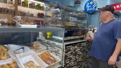 Walter Díaz frente a la vidriera de la panadería La ROU. Viste una camiseta azul manga corta, un pantalón gris y un gorro de visera marrón.