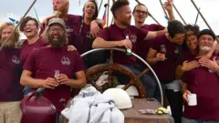 A tripulação do Avontuur sorri para a câmera, segurando bebidas. Eles estão no leme do navio, vestindo camisetas marrons