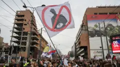 Группа молодых людей протестует с плакатами (в центре плакат с перечеркнутой фигурой) рядом со зданиями, поврежденными бомбардировками.