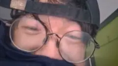 William Li with a blue piece of clothing over his mouth and nostrils. He is wearing a black baseball hat back to front. His glasses are misted up. The walls and ceiling in his flat are behind him.