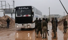 Yağmurlu bir günde çekilen fotoğrafta çamurlu bir yolda bekleyen bir otobüsün önünde silahlı Suriye unsurları var Dokuz erkek görünüyor fotoğrafta ve biri otobüs şöförüyle konuşuyor.