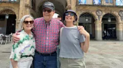 Carlos Montaner abrazado a su esposa, Linda, y su hija, GIna. Los tres sonríen en una plaza.