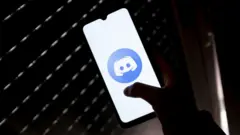 Mão segurando um celular com um logotipo do Discord na tela, em um ambiente escuro