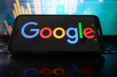 Логотип Google на английском языке с цветными буквами слева направо (синий, красный, от желтого до оранжевого, синего, зеленый, красный) на черном экране смартфона, а на заднем плане приписывается фондовым рынком градиентами синего и зеленого.
