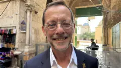 Un hombre con gafas pequeñas de montura metálica redonda, traje gris, camisa blanca y un pequeño distintivo dorado en la solapa sonríe en primer plano. En el fondo se observa una barrera.