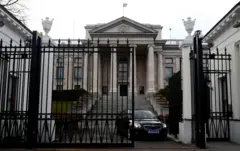 Ambasada Rosji w Warszawie. Duży neoklasyczny budynek, do którego prowadzi ciężka żelazna brama. Na zdjęciu z otwartej bramy wyjeżdża czarny samochód z niebieską tablicą rejestracyjną. 