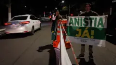 Um homem segura uma faixa escrito 'Anistia já' em um rua, enquanto um táxi passa ao lado. Ao fundo, uma mulher observa com um megafone na mão.