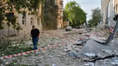 Un hombre pasa junto al lugar de un ataque ruso contra un edificio residencial. Hay escombros en el suelo y una cinta roja y blanca a rayas acordonando la zona. A lo lejos, se ven edificios y árboles. Es de día.