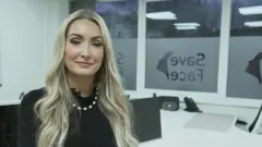 Ashton Collins, long blond hair, black top, white necklace, sit for her white office. Visible for the organization of the organization's organization of the organization of the organization, except the face.