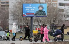Famílias passam, com crianças e carrinhos, caminham em rua em frente a cartaz com foto de militar