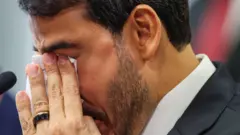 Jorge Messias, de terno, aparece em close, com os olhos fechados e o rosto parcialmente coberto pelas mãos enquanto segura um lenço. Ao lado, há um microfone.