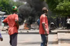 Deux jeunes habillés en rouge observent d'autres jeunes allumant un feu et érigeant un barrage. En arrière plan des jeunes tenant une barrière où un feu est allumé.