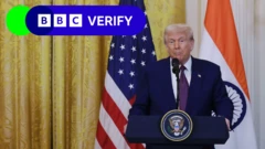 Le président Trump debout devant un rideau doré et les drapeaux de l'Inde et des États-Unis. Il se trouve derrière un pupitre bleu sur lequel figure le sceau présidentiel. Le logo BBC Verify se trouve en haut à gauche de l'image.