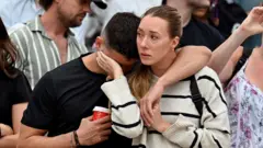 Two people embrace and look away from camera with other mourners behind them, at Bondi Pavilion a day after the shooting.