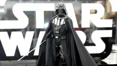 imagen de una persona con un traje de darth vader. tiene el puño cerrado y levantado hacia el frente y en la otra mano sostiene un sable de luz.