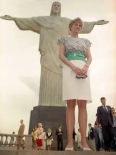 A princesa usa uma saia branca e uma blusa estampada, com um cinco verde. No fundo da foto há várias pessoas.