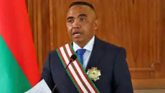 Le colonel Michael Randrianirina lit un discours, vêtu d'un costume, une écharpe sur l'épaule et une étoile de fonction épinglée à sa veste. À côté, on aperçoit le drapeau de Madagascar.
