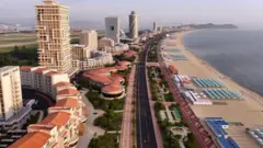 Toma aérea en la que se ven varios edificios, edificaciones más pequeñas, una avenida vacía, algunas zonas verdes, sillas con parasoles muy bien organizadas sobre la arena, en frente al mar