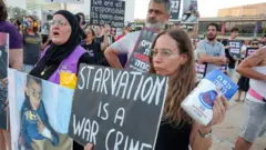 Pessoas com cartazes dizendo que "fome é um crime de guerra" e com imagens de crianças desnutridas