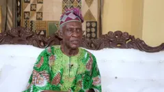 Bíṣọ́ọ̀bù Ladigbolu wọ aṣọ aláwọ̀ ewé tó sì jòkòó lórí àga