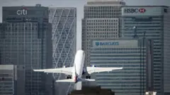 Um avião decola diante de prédios de escritórios com fachada de vidro em Canary Wharf, em Londres, com logotipos visíveis do Citi, HSBC e Barclays