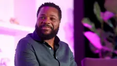 Malcolm-Jamal Warner assis sur la scène en 2023. Il tient un microphone avec le logo de Variety dessus.