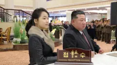 North Korean leader Kim Jong Un (R) and his daughter Kim Ju Ae stand at a hotel reception counter. There are soldiers clapping in the foyer in the background.