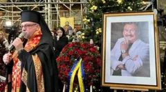 Портрет усміхненого Степана Гіги у білому костюмі на тлі прикрашеної різдвяної ялинки. Поряд – священник із червоним єпитрахилем на грудях і мікрофоном у руці. У тлі – вінки з червоних гвоздик, один з яких із стрічкою з написом "від президента України".