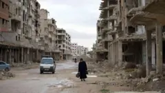 Hasarlı ve yıkık binalarla dolu bir sokaktan geçen bir araba ve elinde çantalarla yürüyen bir kadın