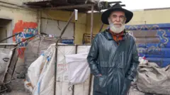 Tahsin Uzun, üzerinde deri ceketi ve kafasında fötr şapkası ile poz veriyor. Arkasında "çekçek" olarak adlandırılan tekerlekli çuvallar var. 