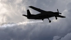 একটি ছোট বিমানের ছায়ামূর্তি দেখা যাচ্ছে, যা আকাশে ছোট ছোট কণা ছিটাচ্ছে এবং তার পেছনে দীর্ঘ সাদা রেখা তৈরি হচ্ছে। বিমানের পেছনে ঘন, ধূসর মেঘ।