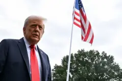 Trump em pé, com olhar sério, ao lado de bandeira dos EUA em área externa