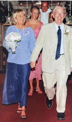Daphne y Chuck saliendo de la iglesia el día de su boda en el Valle de Napa. Daphne, quien sostiene un ramo de flores blancas, lleva una falda azul oscuro hasta los tobillos y un top de manga larga azul claro. Chuck, por su parte, lleva un traje de chaqueta beige, zapatos negros y una corbata azul oscuro a juego con la falda de Daphne.