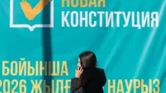 Девушка в темном пальто с непокрытой головой говорит по телефону, проходя мимо плаката, призывающего голосовать на референдуме о новой Конституции Казахстана