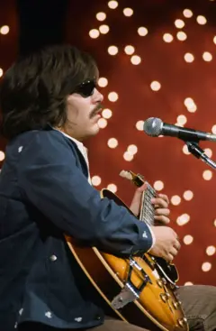 José Feliciano, con su guitarra en mano, sentado frente a un micrófono, con su cabello hasta los hombres y bigote, participa de un especial de NBC. No está claro el año de la foto, pero se le ve de mediana edad.
