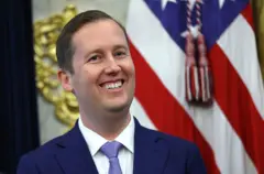 Сержио Гор мовий костюм, оқ куйлак устидан мовий галстук таққан, камерага табассум қилиб қараяпти, орқасида АҚШ байроғи