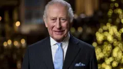 ダークなピンストライプのスーツに淡い青のネクタイとポケットチーフを合わせた国王が笑顔を見せている。背景にはクリスマスツリーの電飾がぼんやりと見える