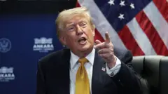Donald Trump hablando y señalando con el dedo. Viste traje oscuro, corbata en tono amarillo-dorado y camisa blanca.