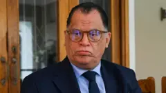 Danny Jordaan, dey wear glasses and dark blue suit wit light blue shirt and dark blue tie