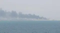 Columnas de humo se elevan desde la derecha de la foto sobre edificios a lo largo de la costa. El mar azul oscuro está en primer plano.