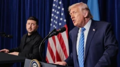 Трамп и Зеленский находятся за трибунами, за ними стоит флаг США. Трамп говорит, Зеленский смотрит на него.